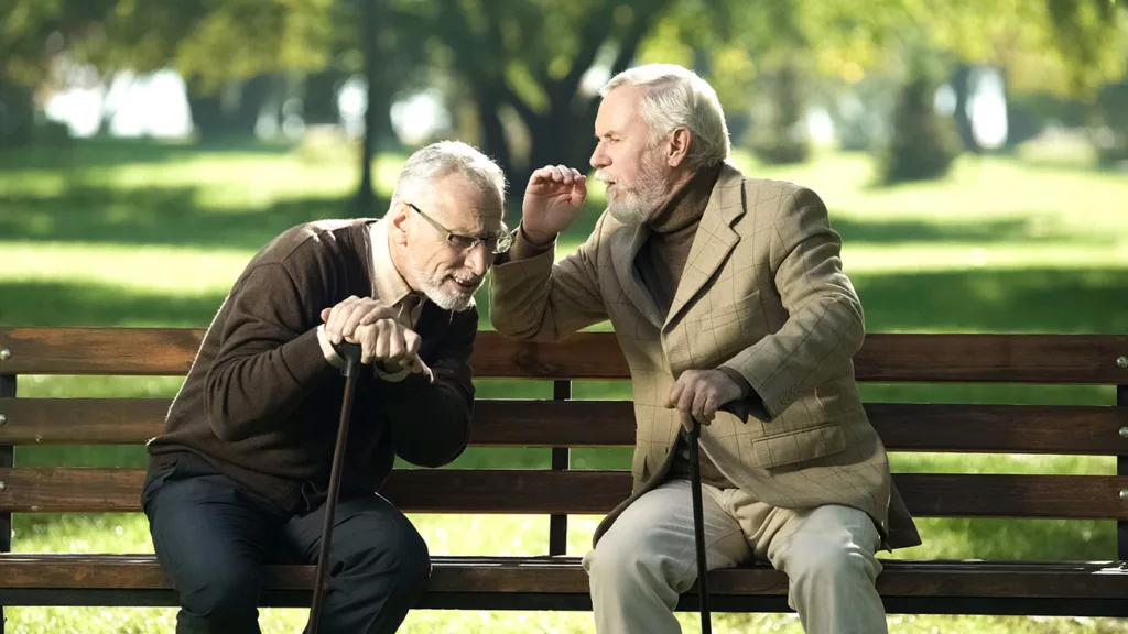 2 older men on park bench with canes. One is leaning in to hear as other puts hand near mouth to speak at him.